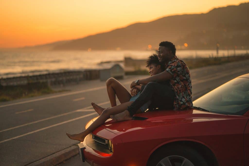 Žena i muškarac promatraju zalazak sunca zagrljeni na haubi auta